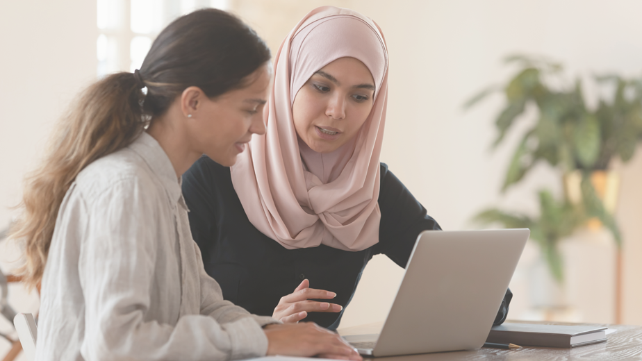 Woman in hijab sitting with smiling colleague at table, looking at computer screen, explaining new support options.
