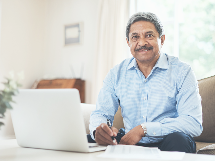 A senior man using a laptop at home while smiling to camera.