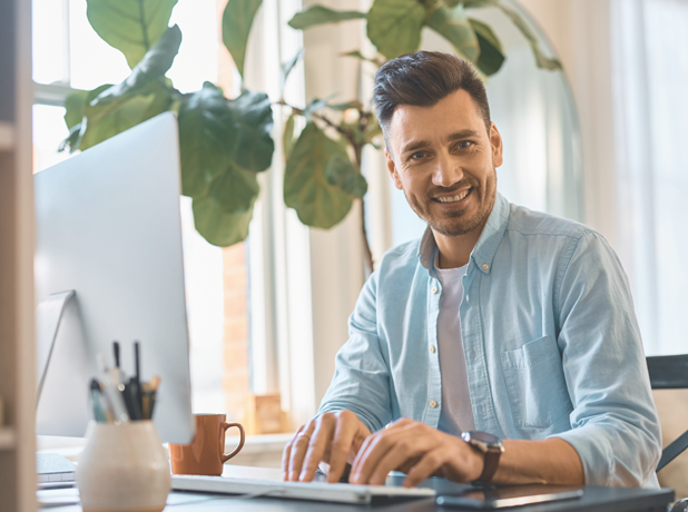 Man at work in a casual office environment working on computer, looking at the camera and smiling.