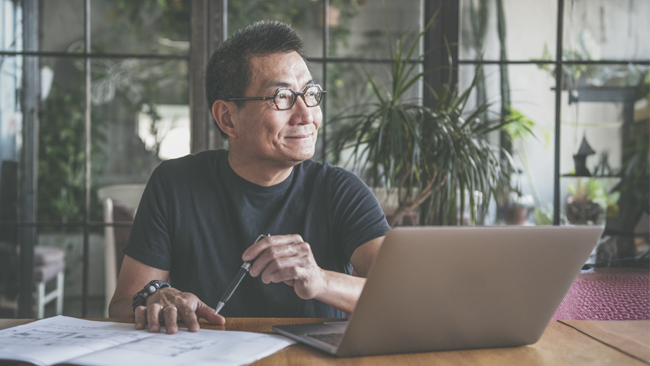 Mature Chinese man using laptop at home looking out the window smiling.