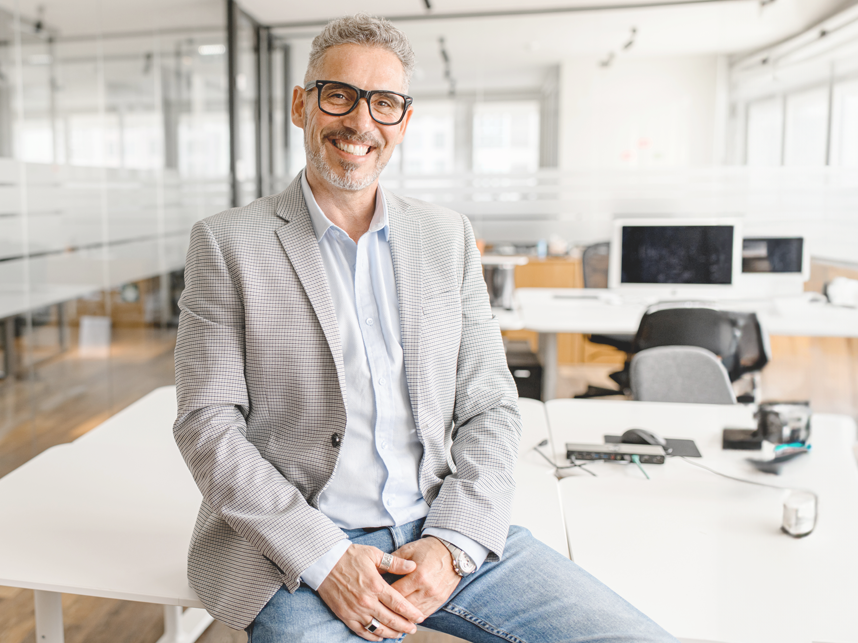 older man in blazer sitting on desk smiling at camera