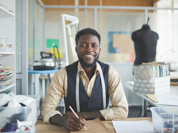 Young cheerful fashion designer sitting by table in his workshop and making new sketches in notepad.