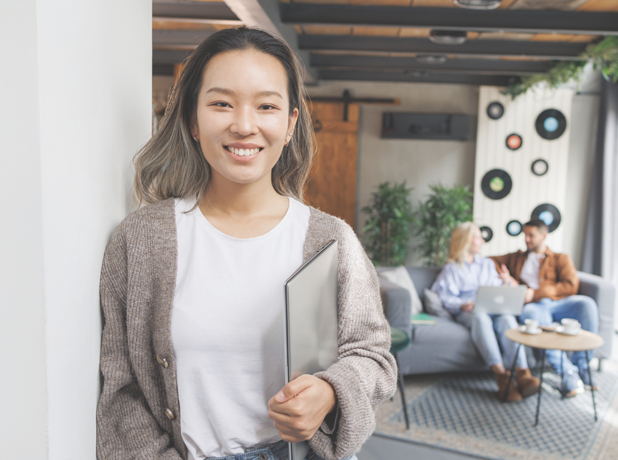 A Chinese woman holding a laptop and looking at the camera in a vibrant modern workplace. Behind her, on the sofa, sits her coworkers, brainstorming and problem-solving, reflecting the dynamic energy of urban productivity.