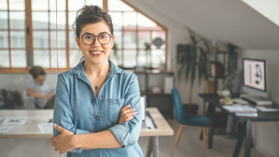 Portrait of young smiling woman in creative office, standing with arms crossed, looking at camera.
