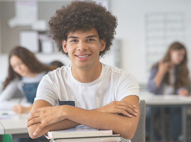 Smiling young student in library leaning on stack of books at his desk with classmates in background. 