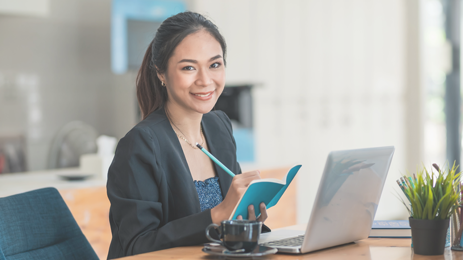 Portrait of a young Asian businesswoman working on a laptop in an office and taking notes in a notebook.