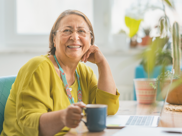 A portrait of a happy hispanic senior woman smiling for the camera while using a laptop at home.