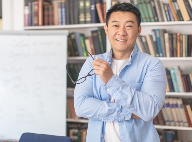 Happy Asian Male Teacher Posing Holding Eyewear Standing In Classroom. 