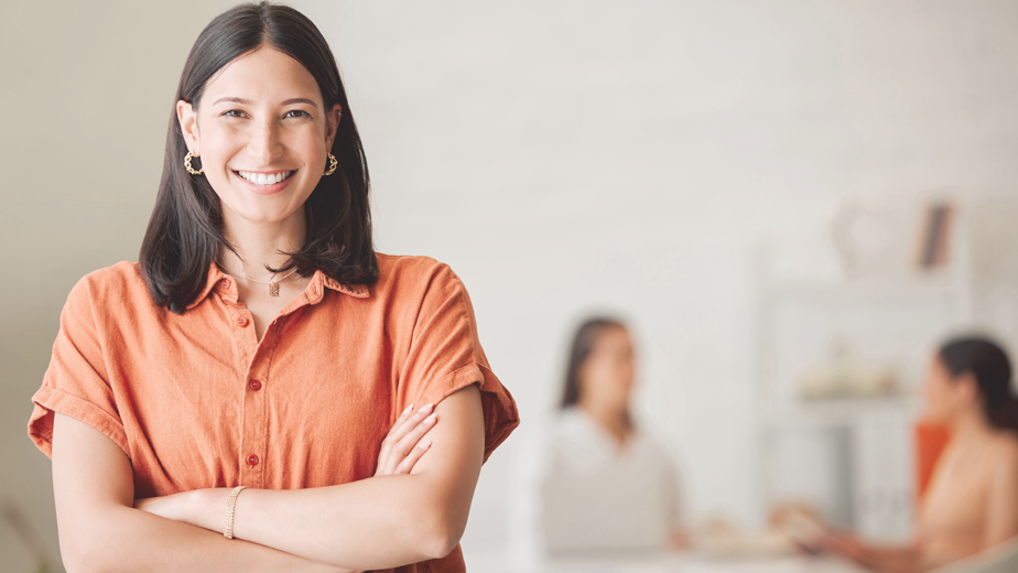 Portrait of one confident young hispanic business woman standing with arms crossed in an office with her colleagues in the background. Ambitious entrepreneur and determined leader ready for success in a creative startup agency.