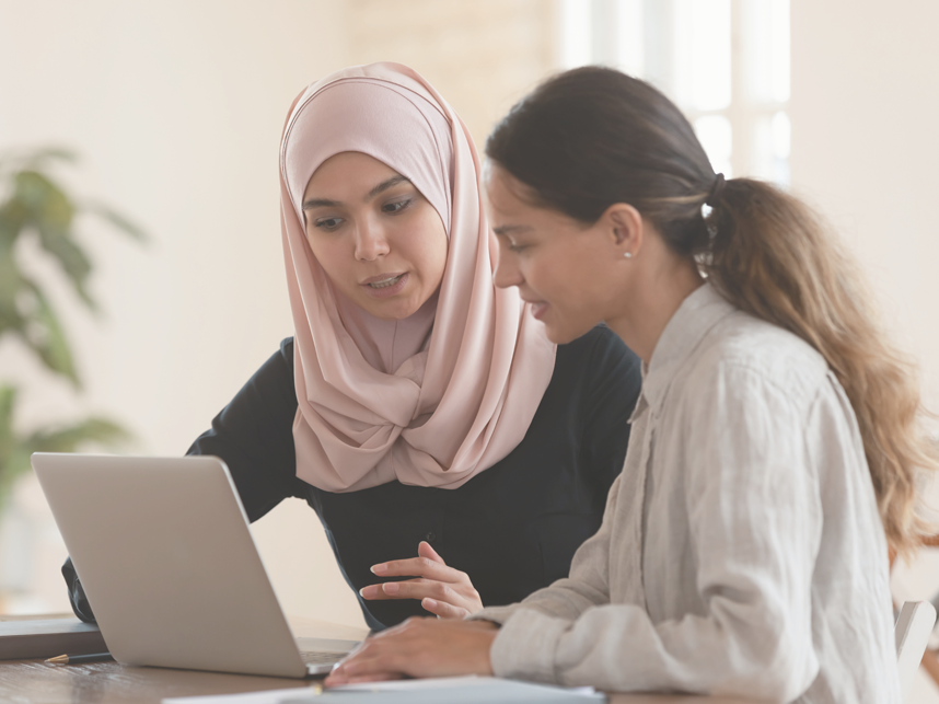 Woman in hijab sitting with smiling colleague at table, looking at computer screen, explaining new support options.