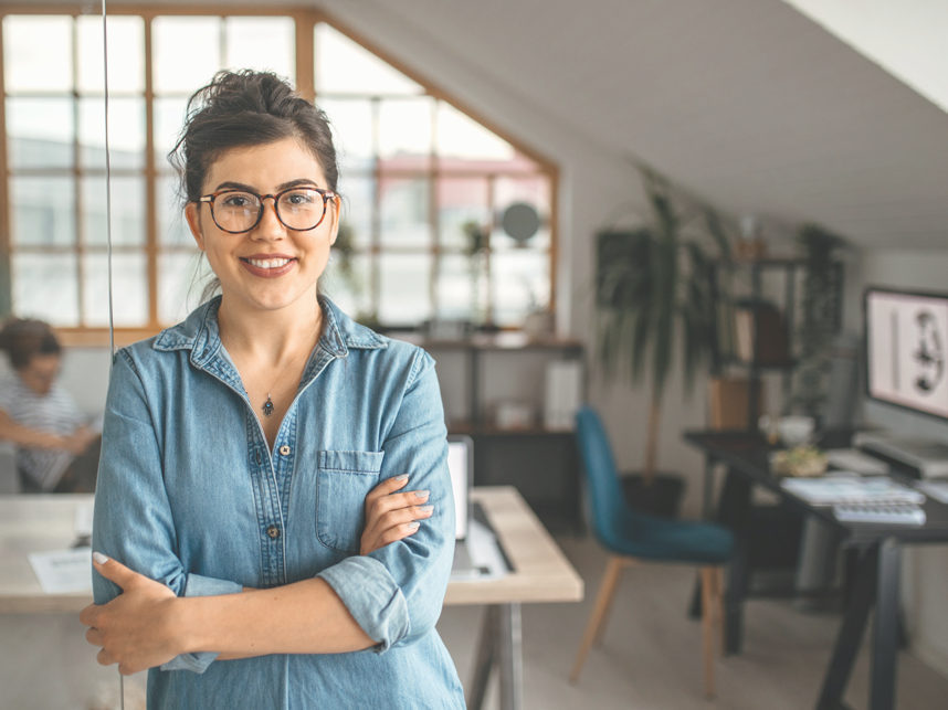 Portrait of young smiling woman in creative office, standing with arms crossed, looking at camera.