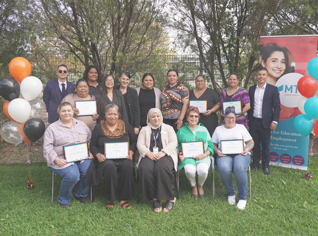 A group shot of MTC Australia's Real Futures Graduation Day event. The group are taking a photo in an outdoor setting with balloons and a branded pull-up banner.