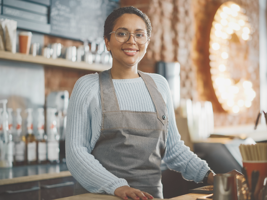 Latin American female barista with short hair and glasses looking at camera and smiling in a coffee shop.