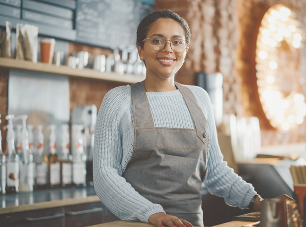 Latin American female barista with short hair and glasses looking at camera and smiling in a coffee shop.