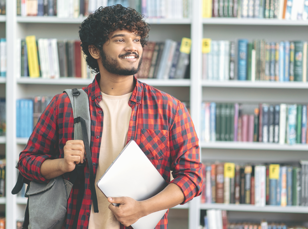 Positive confident Middle-eastern male university student, in stylish casual wear, with backpack and laptop, stands in a library against the background of bookshelves.
