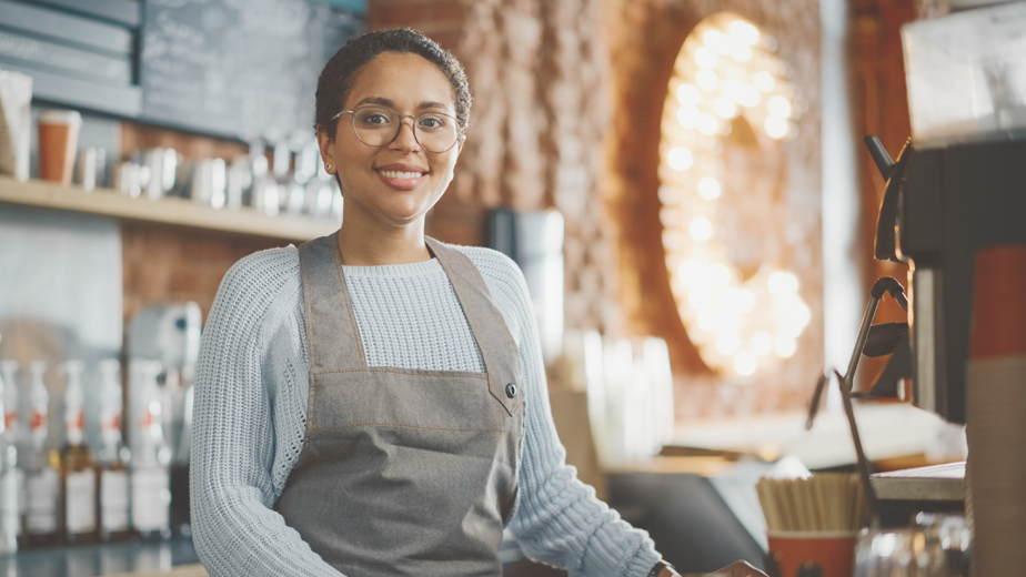 Latin American female barista with short hair and glasses looking at camera and smiling in a coffee shop.
