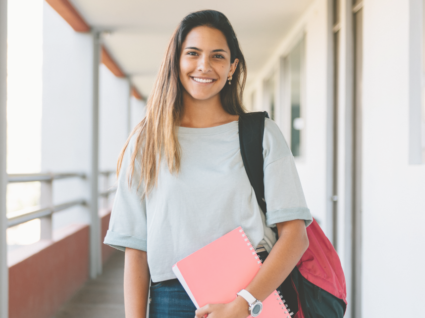 Student on campus looking at the camera and smiling. Carrying a backpack and notebook.