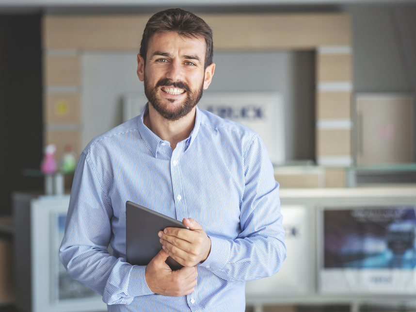 Smiling bearded businessman standing in lobby of shipping firm and holding tablet while looking at camera. 