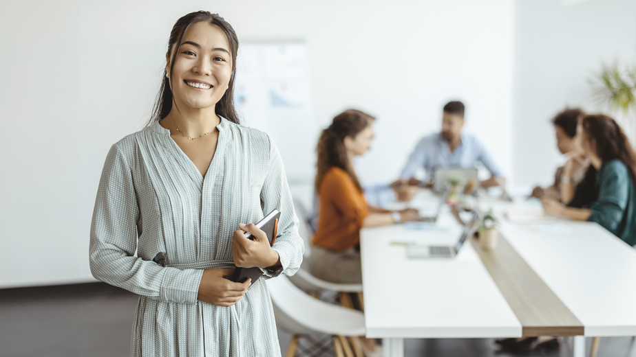 Confident young Asian businesswoman standing smiling at the camera in a boardroom with colleagues in the background.