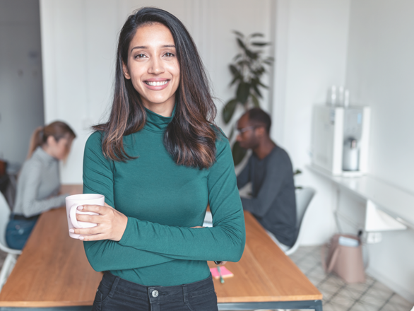 Shot of young indian business woman entrepreneur looking at camera in the office. In the background, her colleagues working.