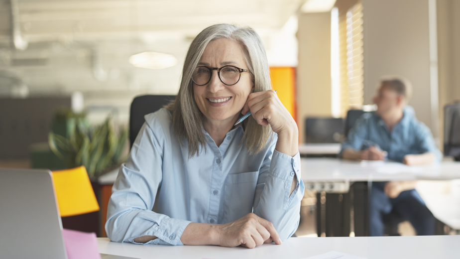 Portrait of smiling confident senior businesswoman, real estate agent, looking at camera in modern office. Gray haired manager wearing stylish eyeglasses sitting at workplace. Successful business