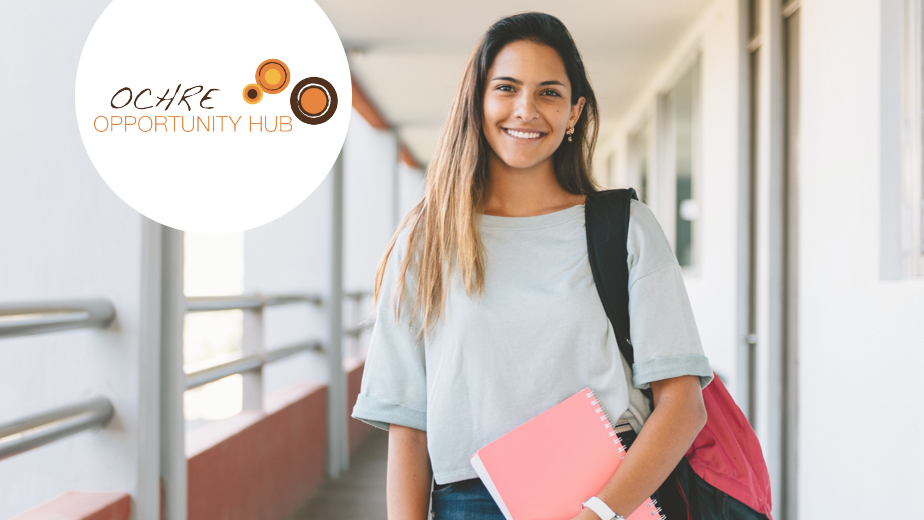 Student on campus looking at the camera and smiling. Carrying a backpack and notebook.