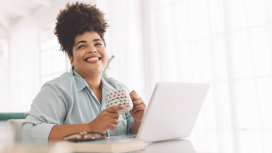 Wide angle shot of happy African American  woman sitting on table with coffee mug and laptop while working from home. 