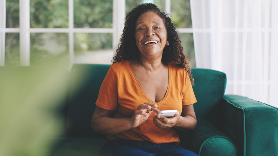 An Islander woman in orange sits on her couch smiling and laughing at the camera as she types on her phone.