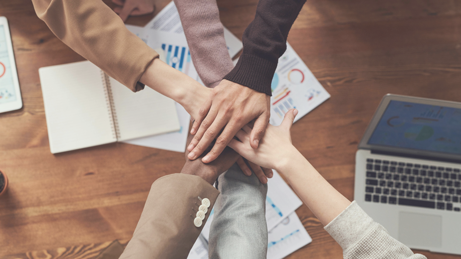 A team of people hold their hands stacked on top of each other above their work table filled with papers