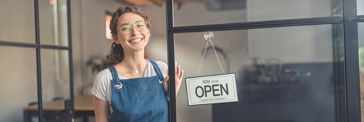 Youn,g joyful, curly-haired waitress with glasses standing at cafe entrance next to open for business sign. 