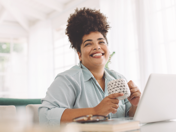 Wide angle shot of happy African American  woman sitting on table with coffee mug and laptop while working from home. 