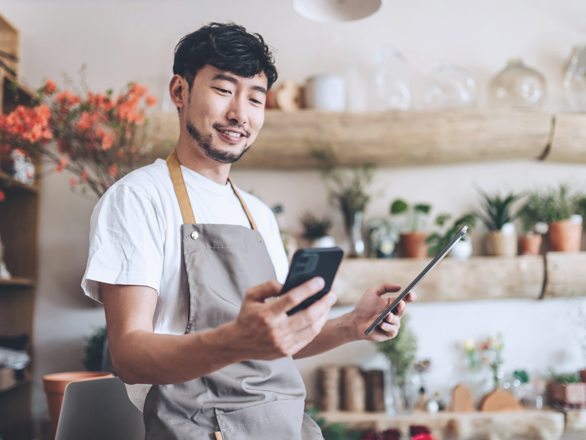 Asian male florist, owner of small business flower shop, using smartphone while working on digital tablet in work place. Checking stocks, taking customer orders, selling products online. Daily routine of running a small business with technology