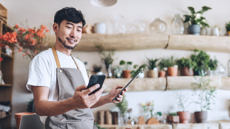 Asian male florist, owner of small business flower shop, using smartphone while working on digital tablet in work place. Checking stocks, taking customer orders, selling products online. Daily routine of running a small business with technology