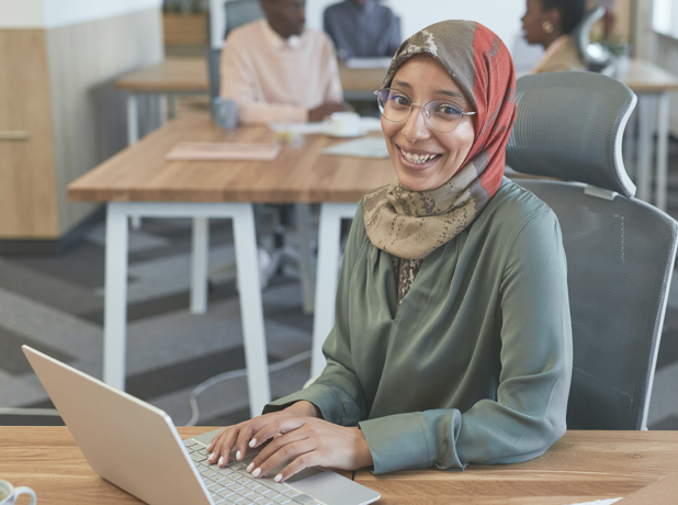 Smiling woman in headscarf and glasses sitting at desk on laptop