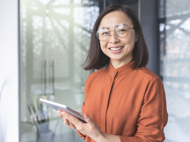 Portrait of a young Asian female with glasses smiling confidently to camera holding a touch screen device.