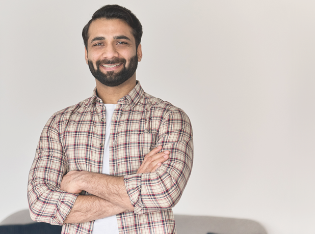 Portrait of young happy indian business man looking at camera standing with arms crossed at home office.