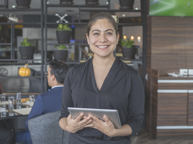 Hispanic waitress standing and smiling to camera holding touchpad at a high-class restaurant.