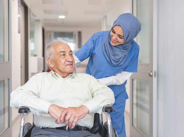 Female, Muslim aged-care worker smiling and looking down at elderly man whilst pushing his wheelchair.