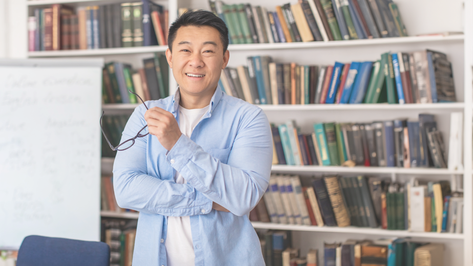 Mature asian man holding glasses in library smiling at camera.