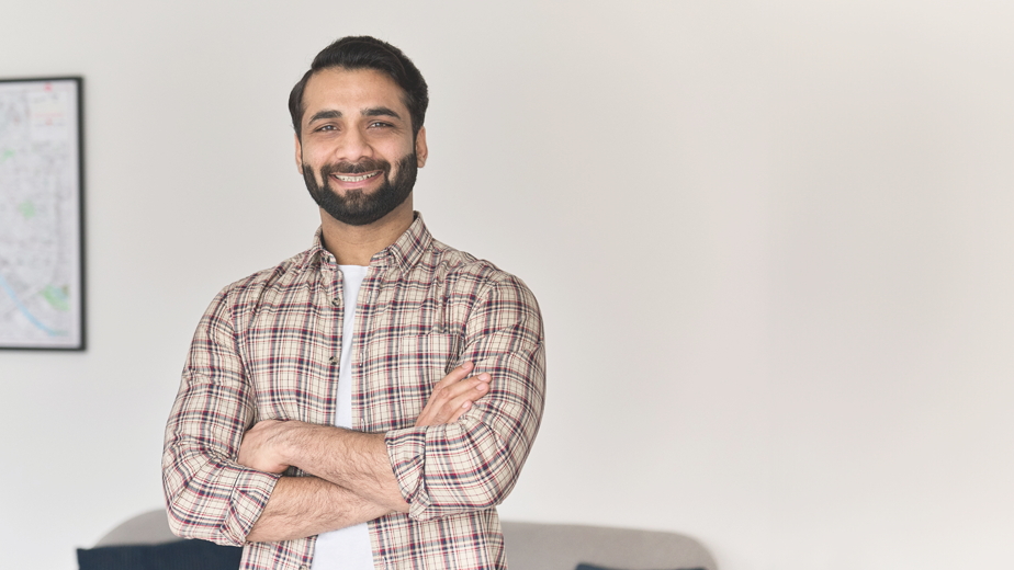 Portrait of young happy indian business man looking at camera standing with arms crossed at home office.