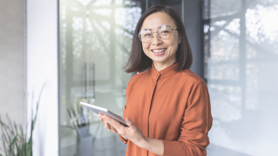 Portrait of a young Asian female with glasses smiling confidently to camera holding a touch screen device.