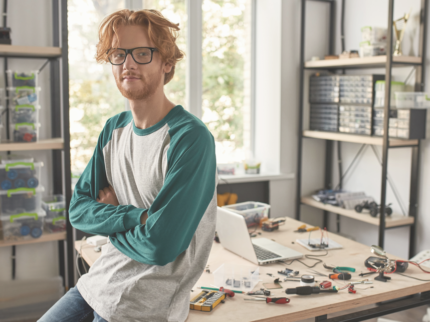 Male red haired Caucasian technician leaning on desk with crossed arms looking at camera in office. There are a lot of tools and equipment on the desk.