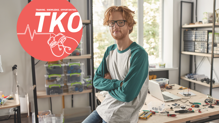 Male red haired caucasian technician leaning on desk with crossed arms looking at camera in office. There are a lot of tools and equipment on the desk.