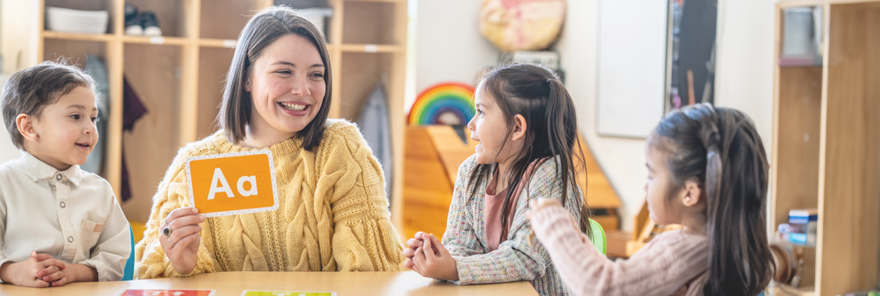 A Kindergarten teacher sits with a small group of students as she teaches them the different sounds each letter of the alphabet makes. Each of the students are dressed casually and paying close attention to their teacher.