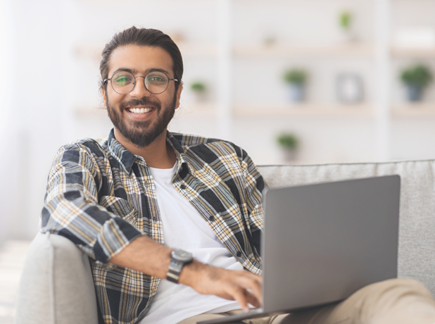 Positive Indian male reclining on couch with laptop whilst completing online study.